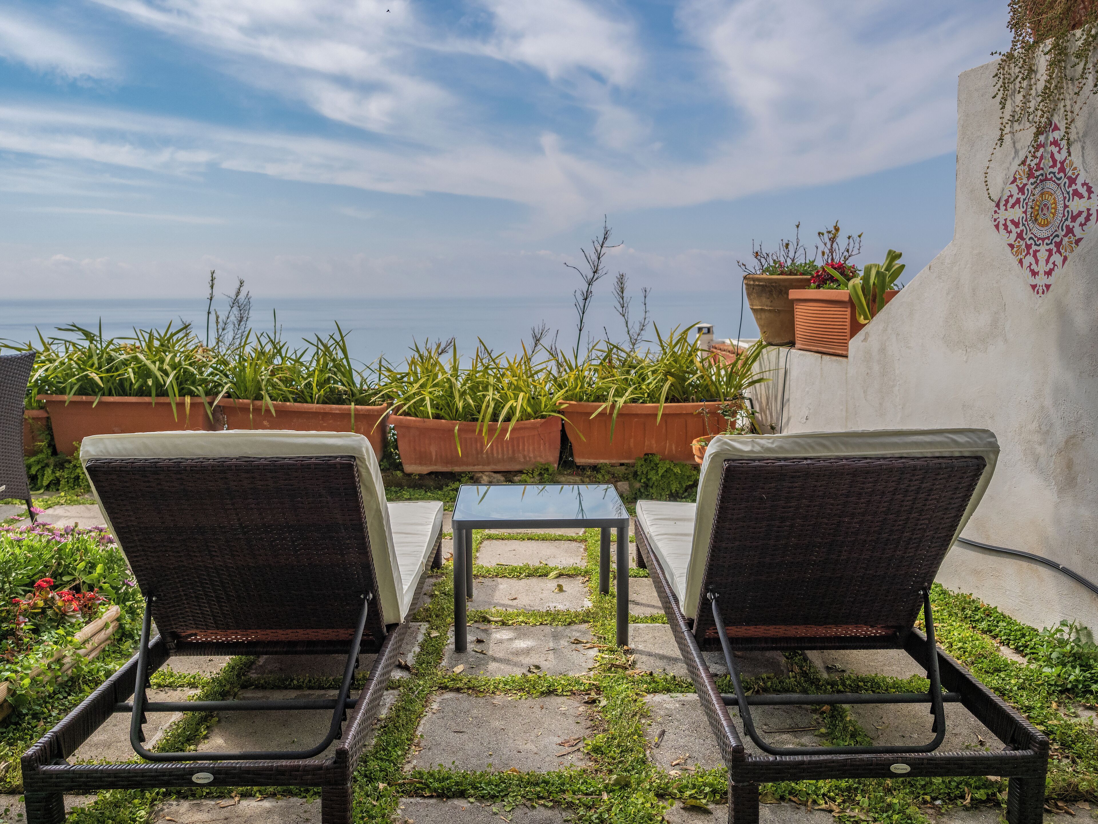 Two wicker lounge chairs facing a view of the sea, with potted plants and a patterned wall.