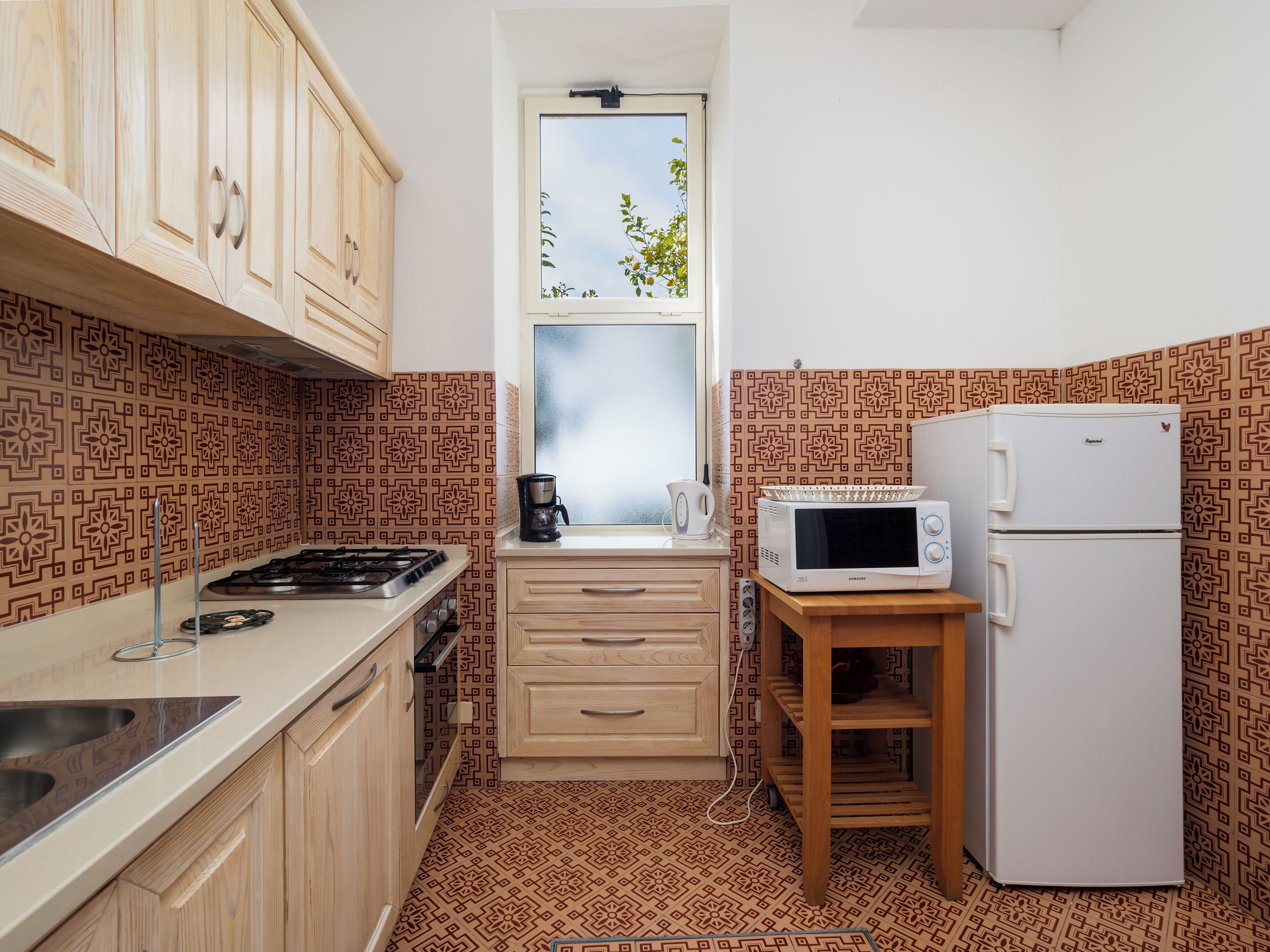 A compact kitchen with a gas stove, a microwave, a refrigerator, and patterned tiles.