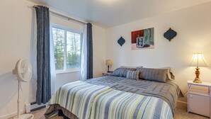 Quarto com uma cama grande, um ventilador, janela com cortinas, mesa de cabeceira com abajur e um quadro na parede.