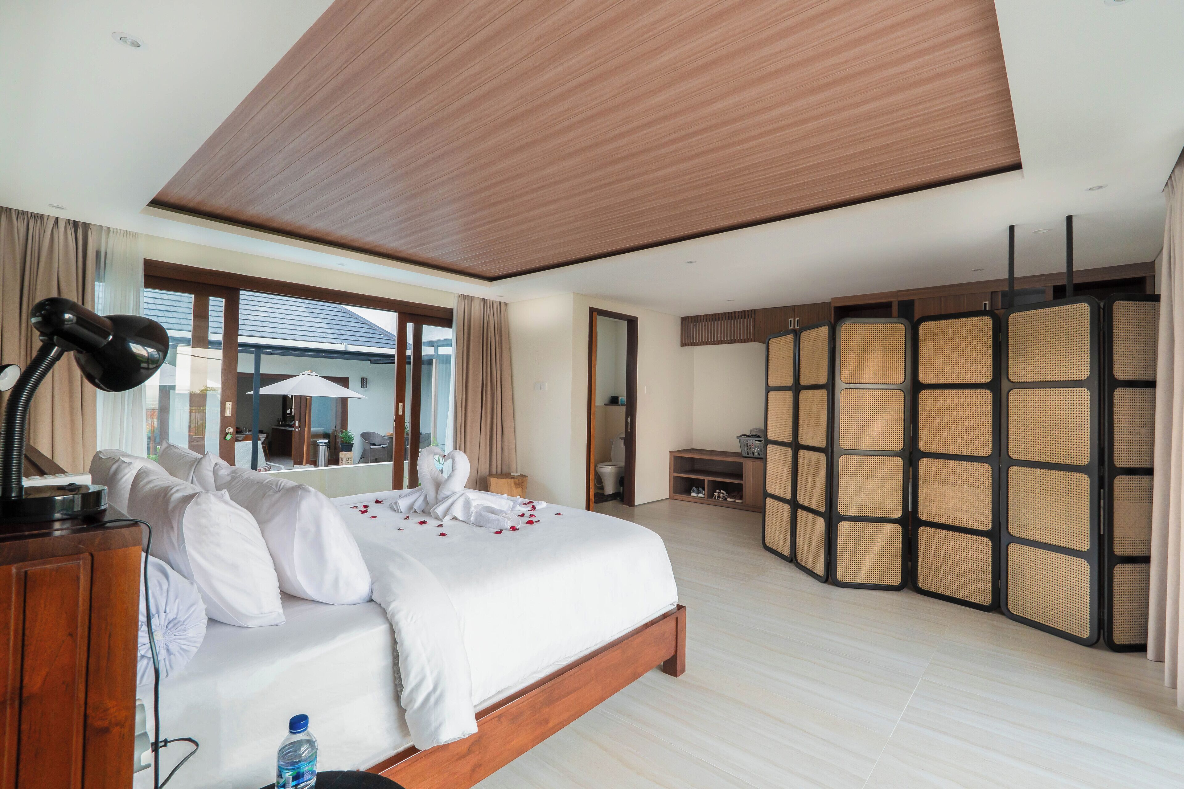 A hotel room with a large bed, wooden headboard, a desk, a mirror, and a partition with a woven design.