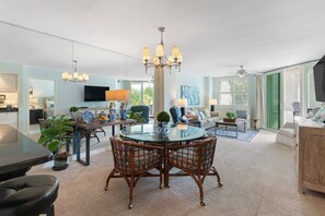A spacious living room with a glass dining table, wicker chairs, a chandelier, and a flat-screen TV.