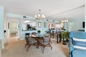 A modern living room with a round glass table, wicker chairs, a blue sofa with 'welcome' pillow, and a kitchen in the background.