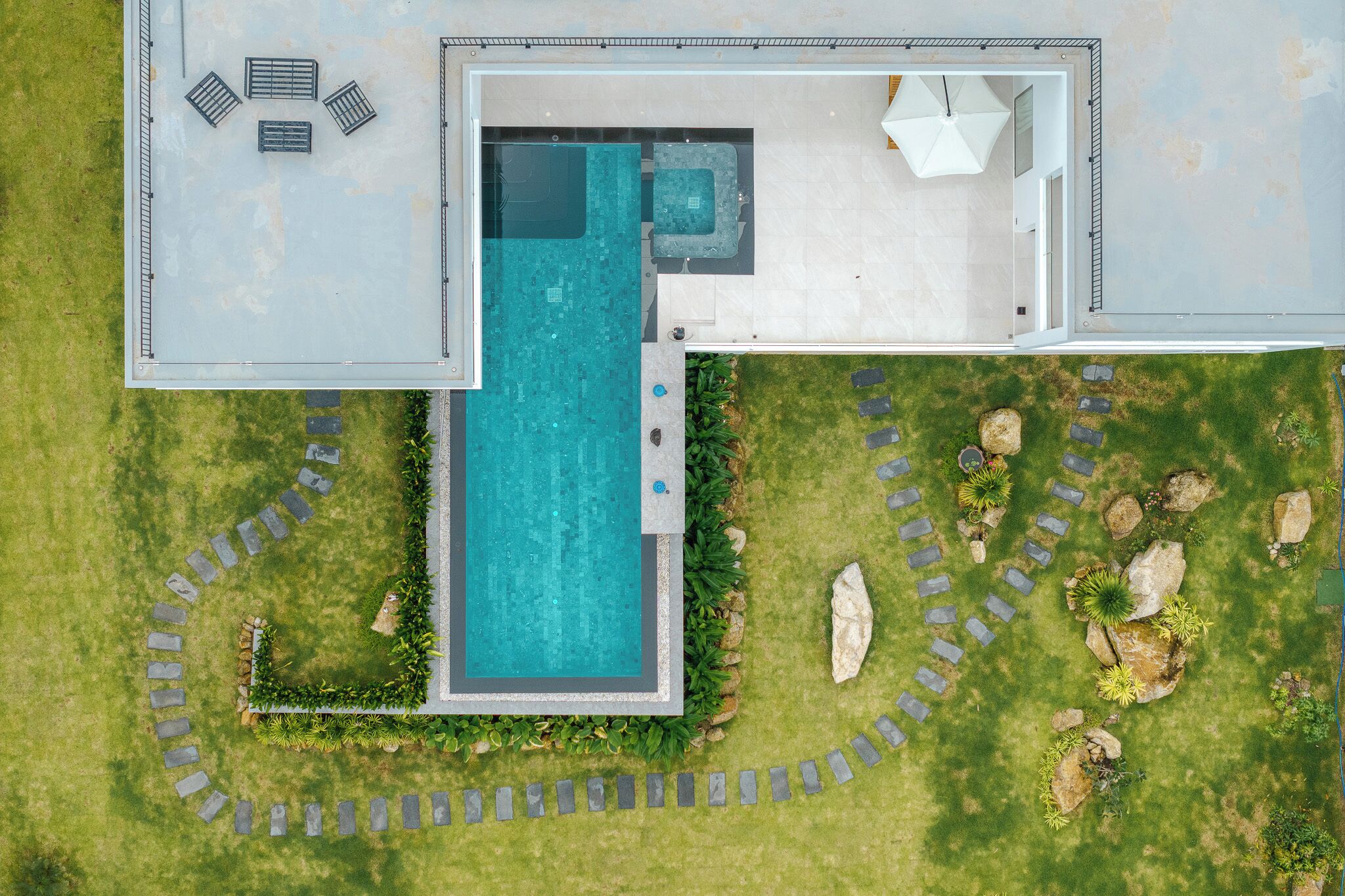 An aerial view of a modern house with a rectangular swimming pool, a patio area with seating, and a well-maintained garden with a stone pathway.