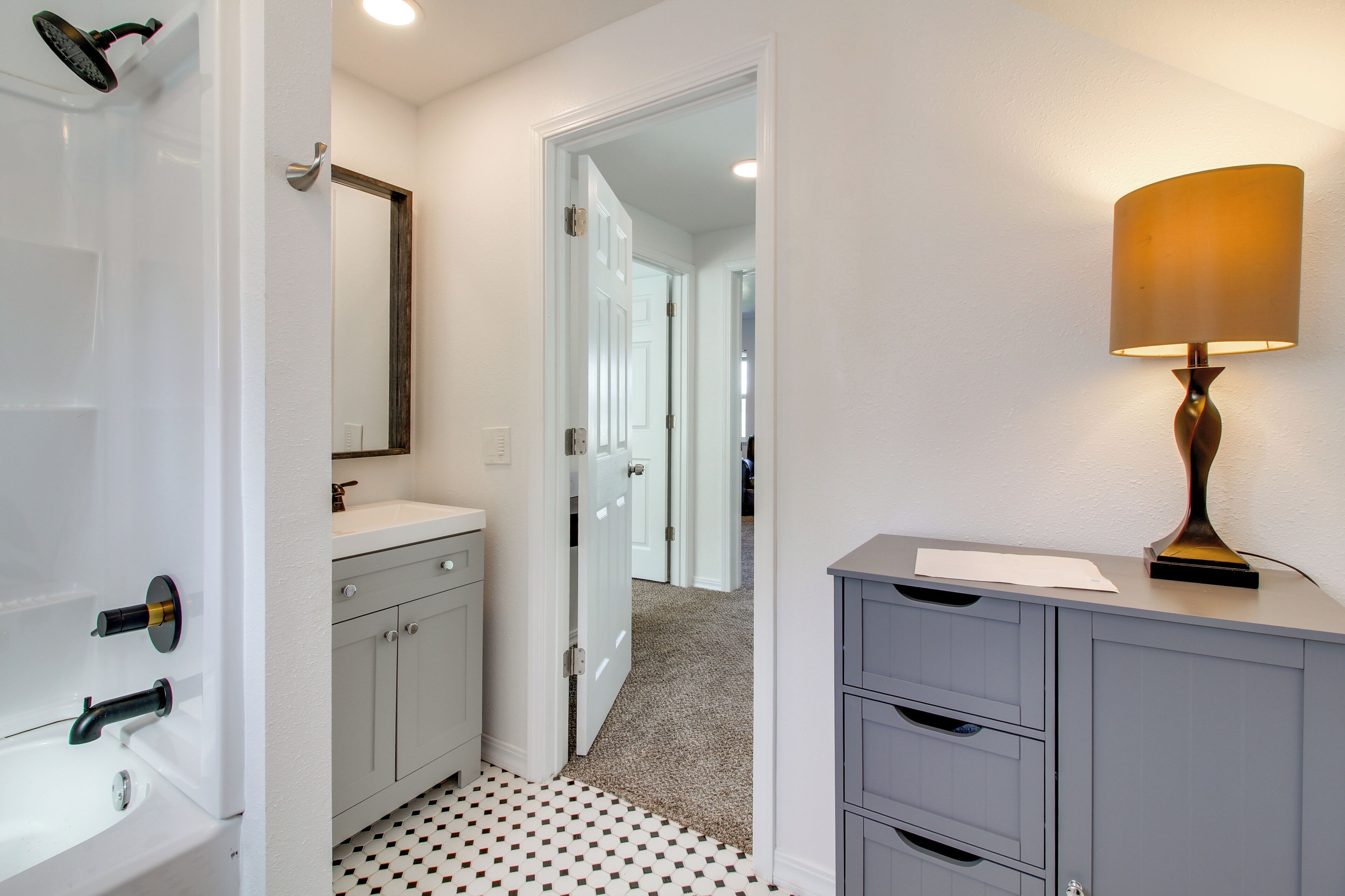 A bathroom with a white sink, a mirror, and a shower. There is a cabinet with drawers and a lamp on top.
