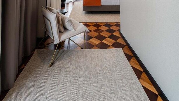 Une chambre d’hôtel avec un sol à carreaux, un lit recouvert d’une couverture orange, une chaise, un tableau abstrait encadré et une vue sur une autre chambre avec un lit et des rideaux.