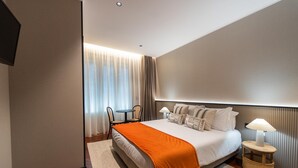 A modern hotel room with a large bed, a small table with chairs, a desk, and a window with curtains.