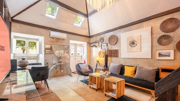 A spacious living area with a glass coffee table, a sofa with cushions, a black chair, and a wooden side table. There is a fireplace with a decorative vase on the table, and a large window providing natural light. - GuestReady - Classic prestige in Costa da Caparica (Costa da Caparica)