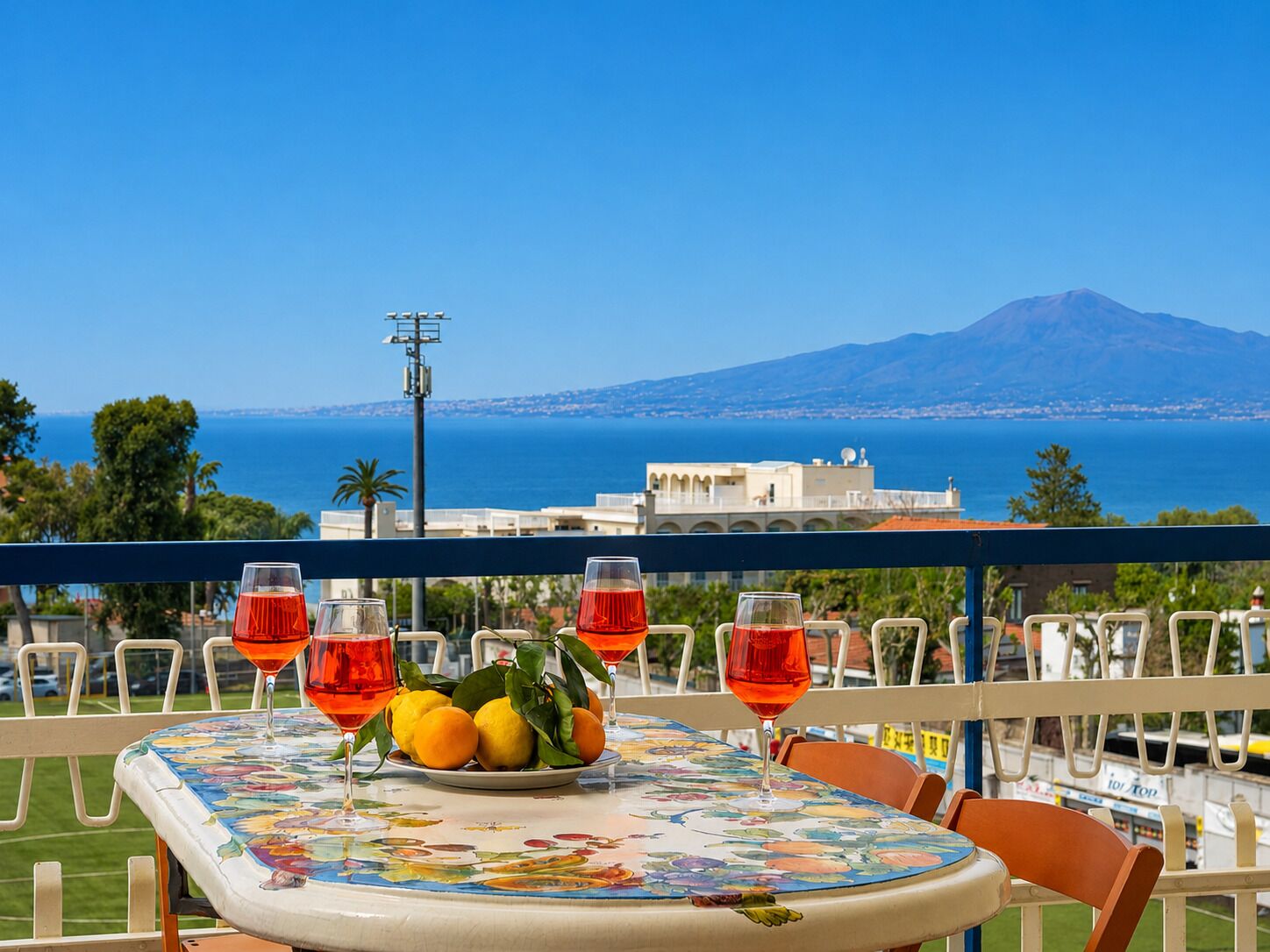 A table with a floral tablecloth, a plate of lemons, and four glasses of red drink, overlooking a coastal view with a mountain in the distance.