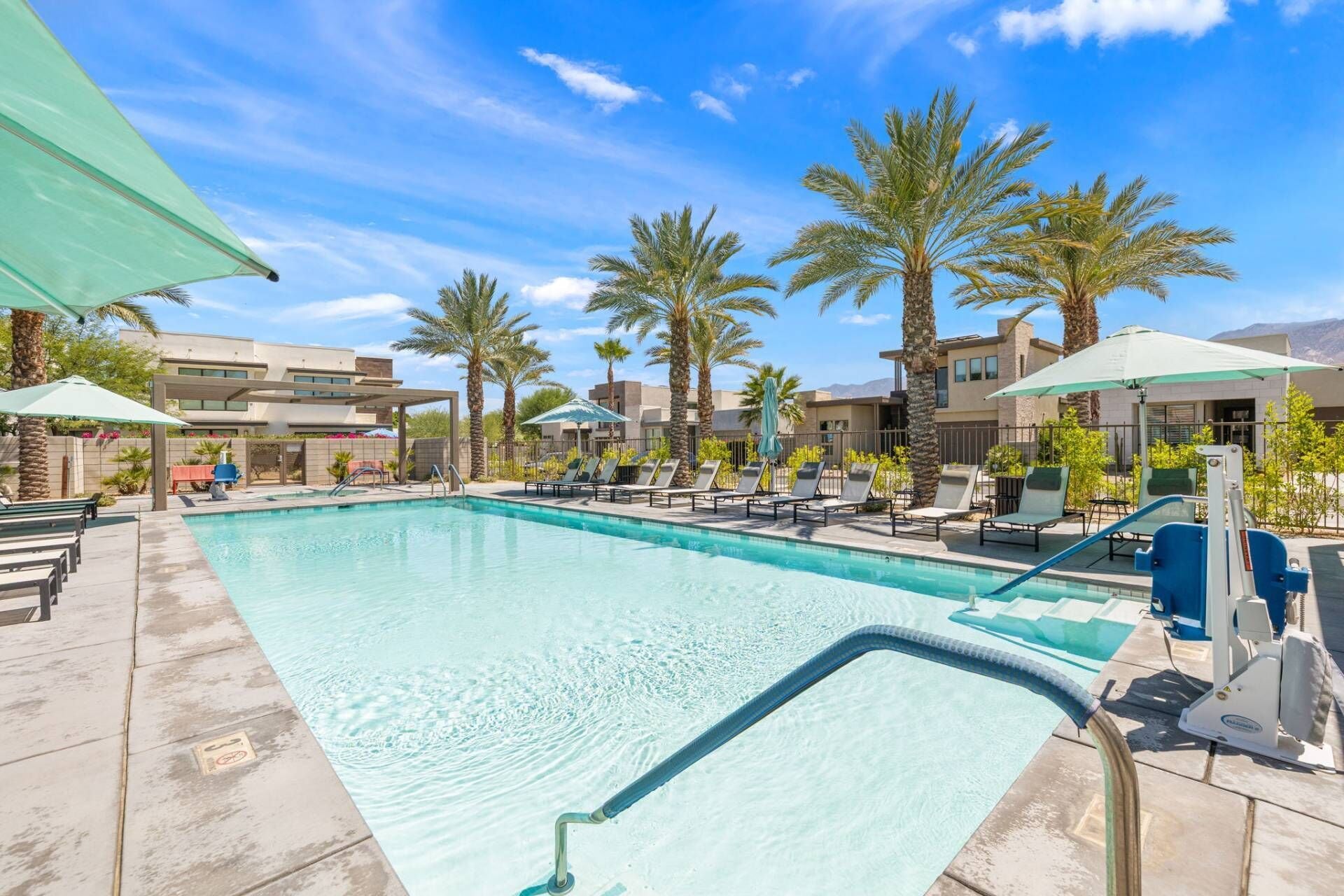 A swimming pool with lounge chairs and umbrellas, surrounded by palm trees and modern buildings.
