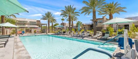 A swimming pool with lounge chairs and umbrellas, surrounded by palm trees and modern buildings.