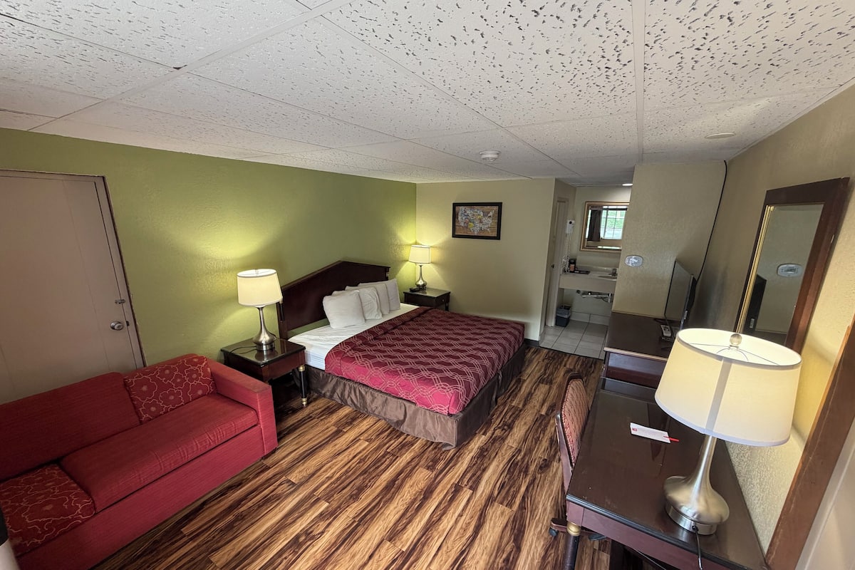 Une chambre d’hôtel avec un lit, un canapé rouge, un bureau avec une lampe, un miroir et une salle de bain visible à travers une porte ouverte.