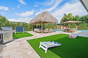 Un jardin avec un barbecue, un gazebo à toit de chaume, une piscine et une table pour jouer à des jeux de plein air.