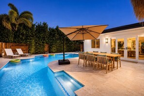 Une piscine avec des chaises longues, une table avec des chaises et un grand parasol.