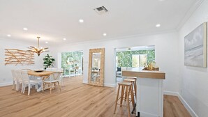 Ein geräumiger Raum mit Holzboden, weißen Wänden, einem Essbereich mit weißen Stühlen, einer Küchenzeile mit weißer Arbeitsplatte und einem großen Fenster mit Blick auf Grünflächen.