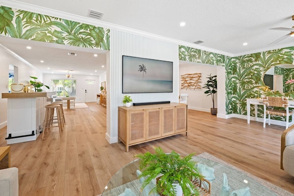 A modern living room with a flat-screen TV, wooden flooring, and tropical wallpaper. - Spacious Designer 4BR Pool Home Near Beach | BBQ & Games (Fort Lauderdale)
