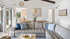 Ein modernes Wohnzimmer mit einem grauen Ecksofa, einem Esstisch mit Stühlen, einer Küche im Hintergrund und einer großen Wanduhr.