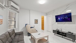 Een moderne woonkamer met een grijze bank, een massagestoel, een houten salontafel, een televisie bevestigd aan een marmeren muur, en een console met een gameconsole.