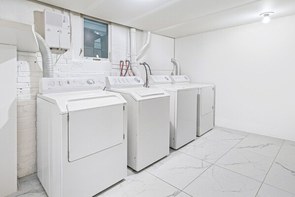 Une buanderie équipée de trois machines à laver blanches et d’un sèche-linge, ainsi que d’un système de ventilation et d’une fenêtre.