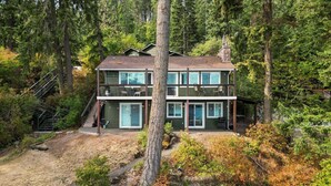 A two-story cabin with a large deck, surrounded by dense forest.