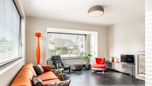 Un salon moderne avec un canapé, des fauteuils, un téléviseur et une grande fenêtre équipée de stores.