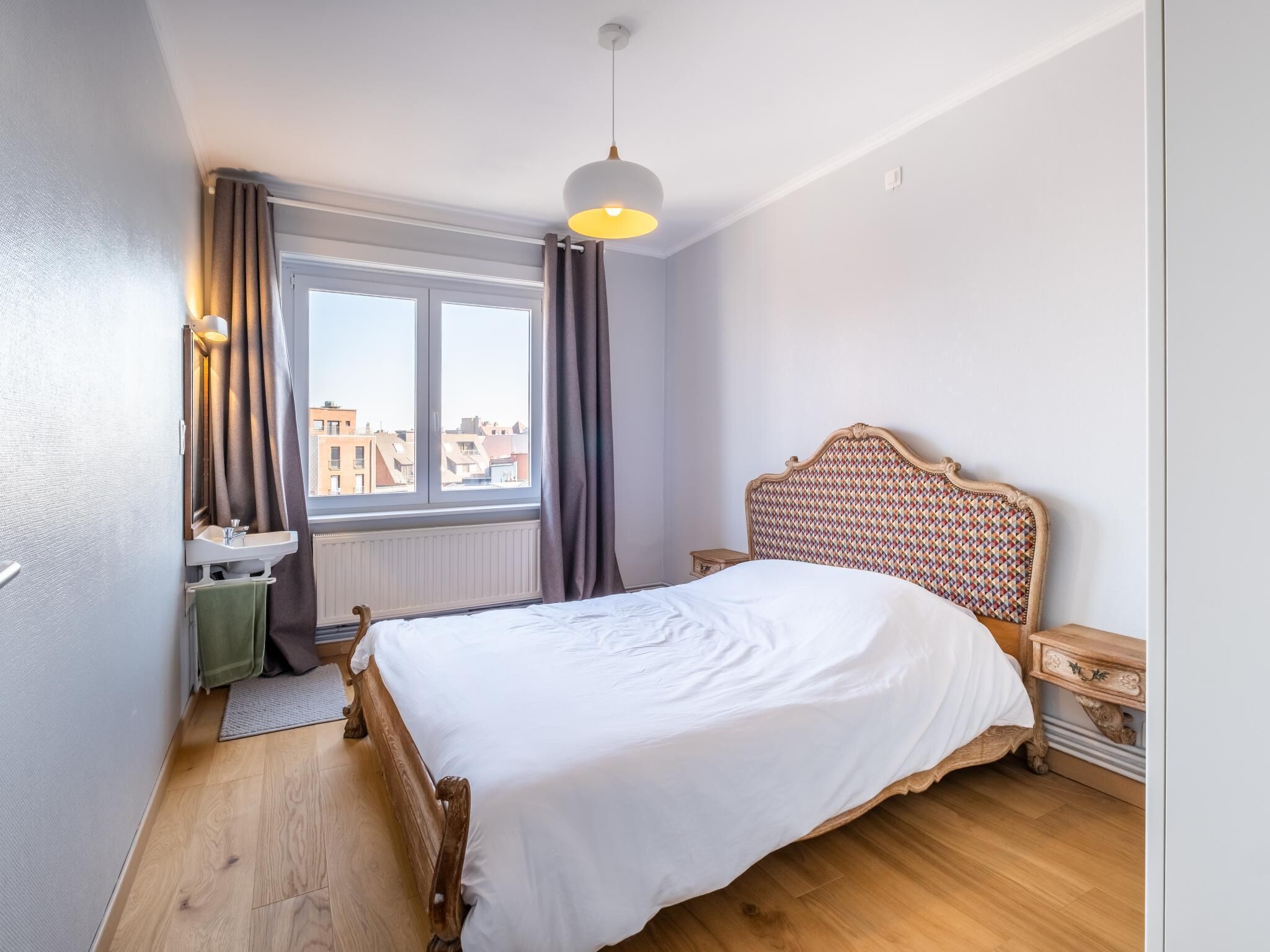 A bedroom with a wooden bed, a window with curtains, a bedside table, and a hanging light fixture.