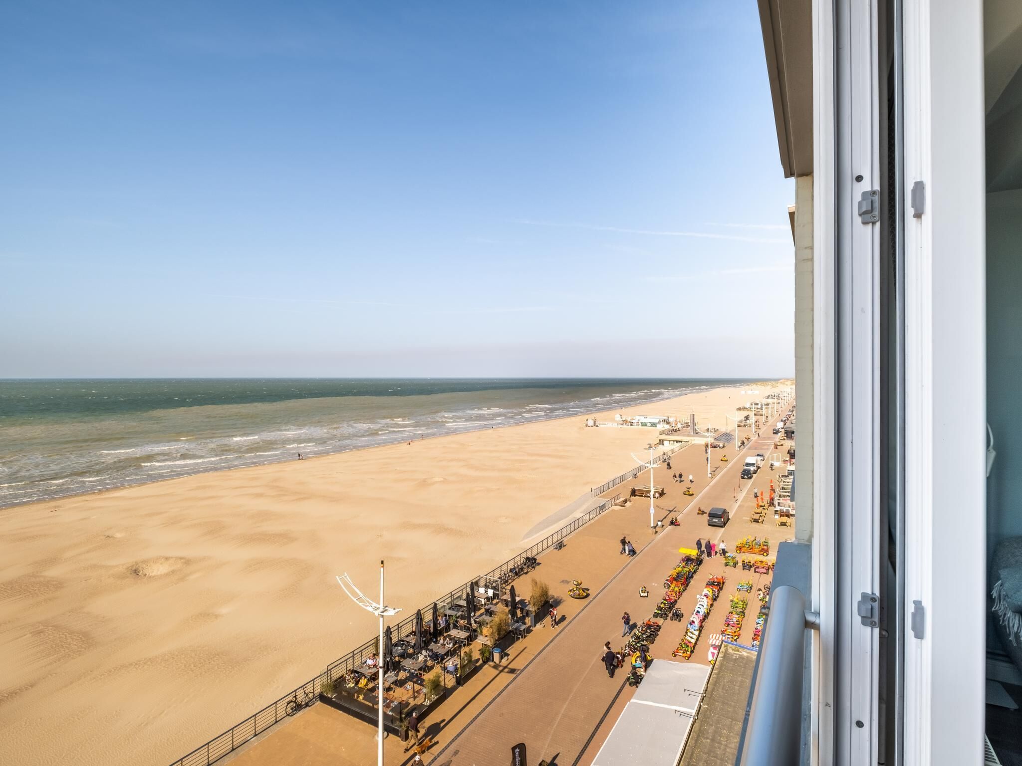 Blick vom Balkon auf einen Sandstrand mit Promenade, Geschäften und wolkenlosem Himmel.
