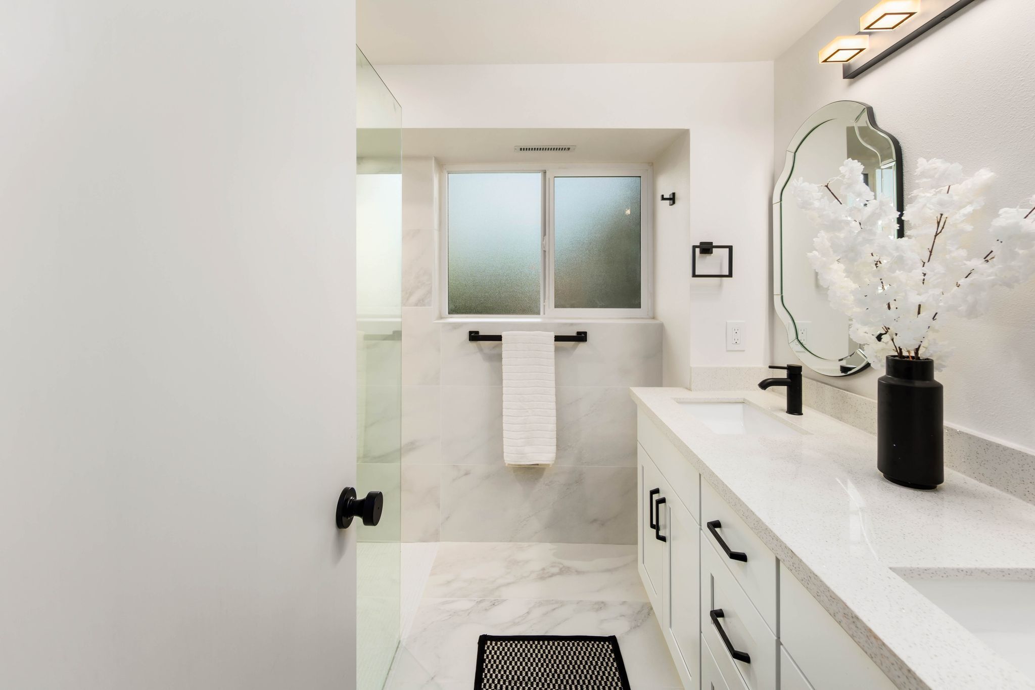 A modern bathroom with a glass shower enclosure, a white marble floor, a white vanity with black handles, and a large mirror with a vase of white flowers.