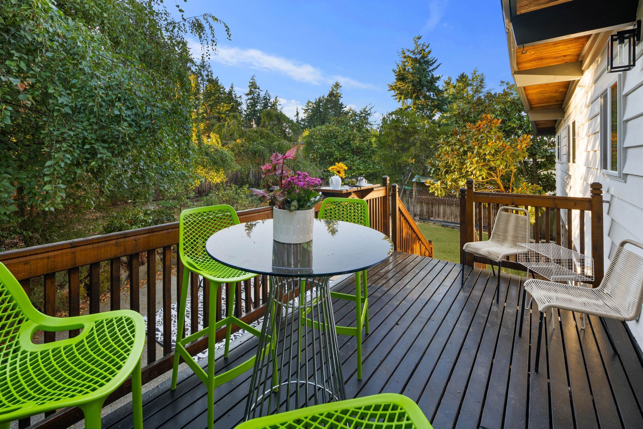 Una terraza de madera con una mesa redonda de vidrio y sillas verdes, rodeada de exuberante follaje y árboles.