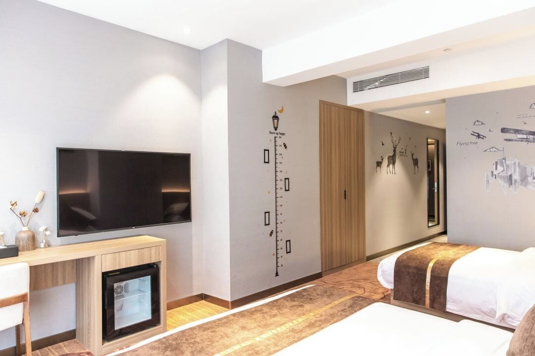 A modern hotel room with a flat-screen TV, a mini-fridge, a bed with a headboard, a wall with a height chart, and a mirror.