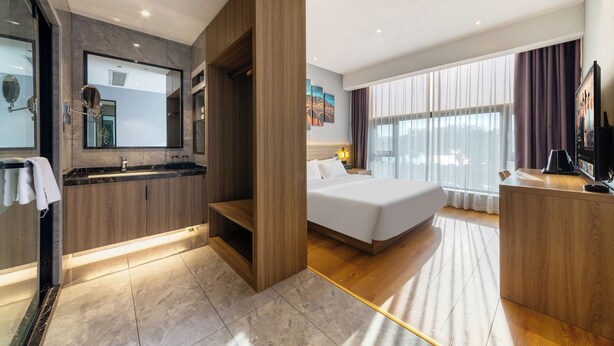 Une chambre d’hôtel moderne dotée d’un grand lit, d’un bureau en bois, d’une salle de bain avec un miroir et un lavabo, et d’une grande fenêtre avec des rideaux.
