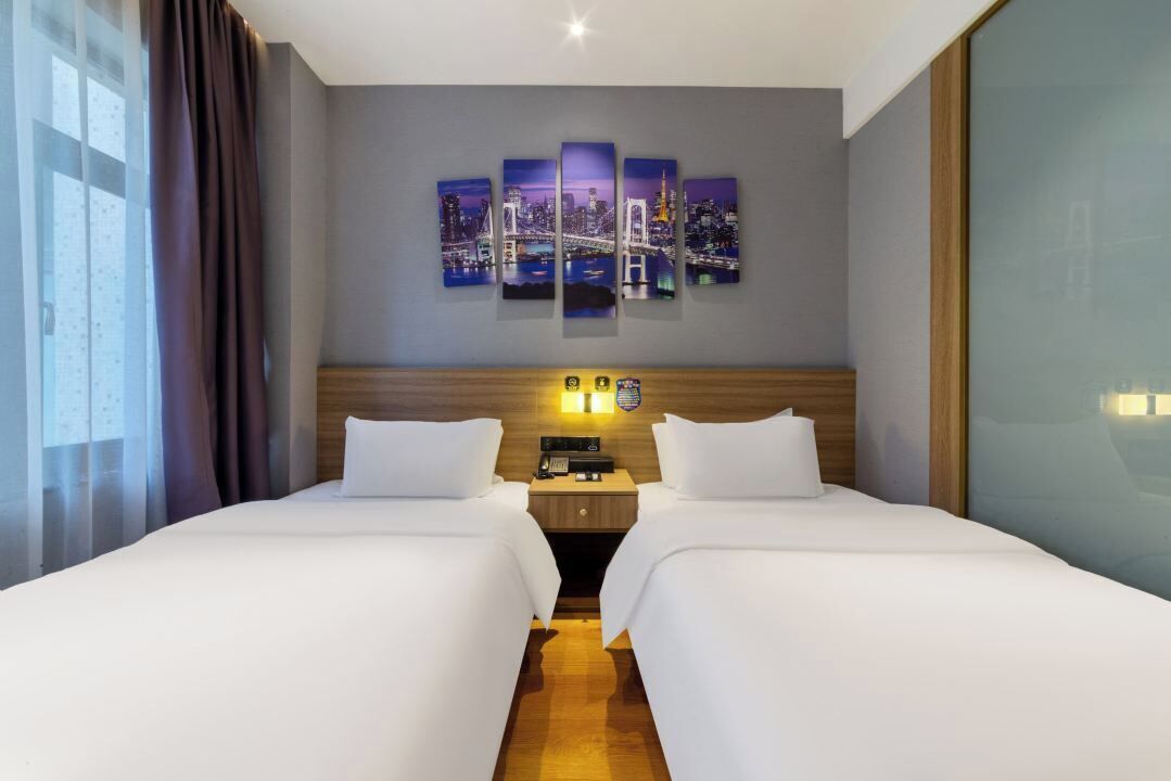 A hotel room with two beds, a cityscape artwork on the wall, a nightstand with a phone and lamp, and a window with curtains.