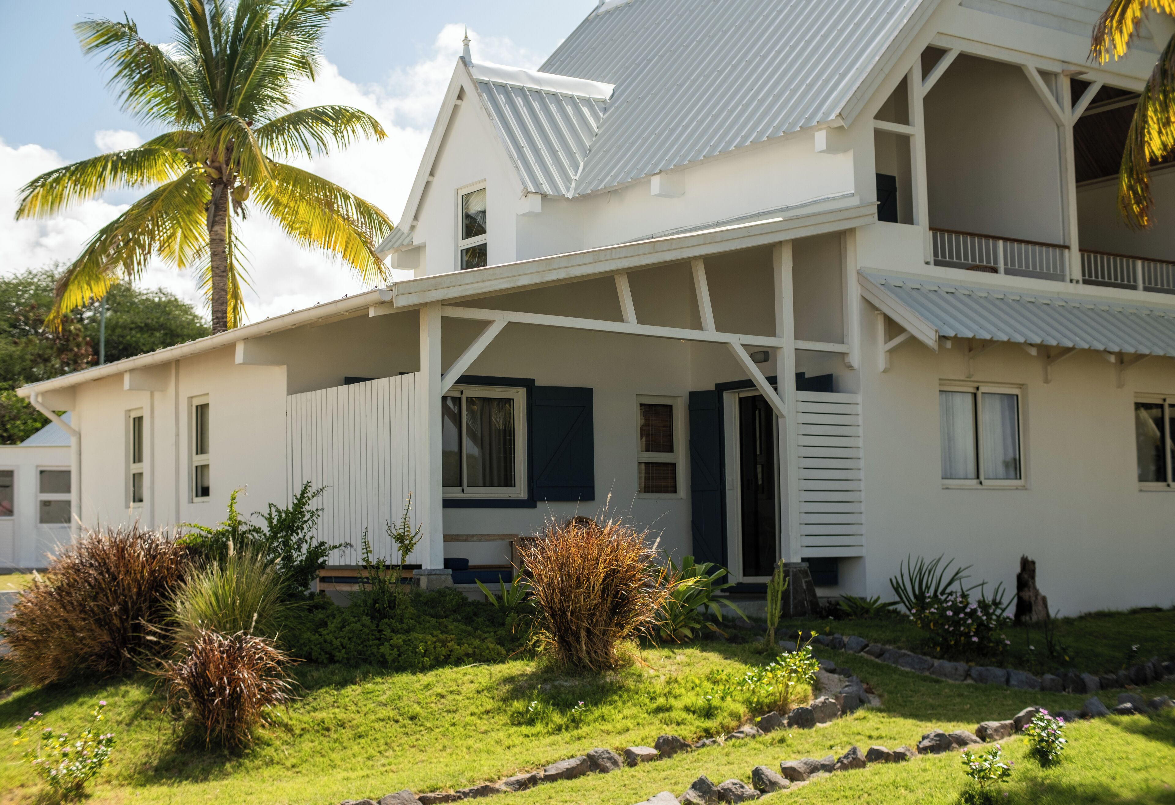 Una casa de estilo colonial blanco con un porche cubierto, una palmera y un jardín bien cuidado.