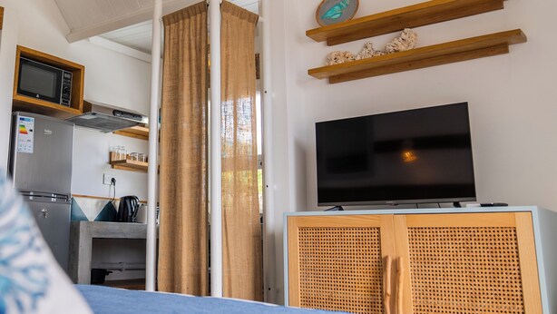 Une chambre avec un lit, un téléviseur posé sur un meuble en bois, une kitchenette équipée d’un four à micro-ondes et d’un réfrigérateur, et une fenêtre avec des rideaux.
