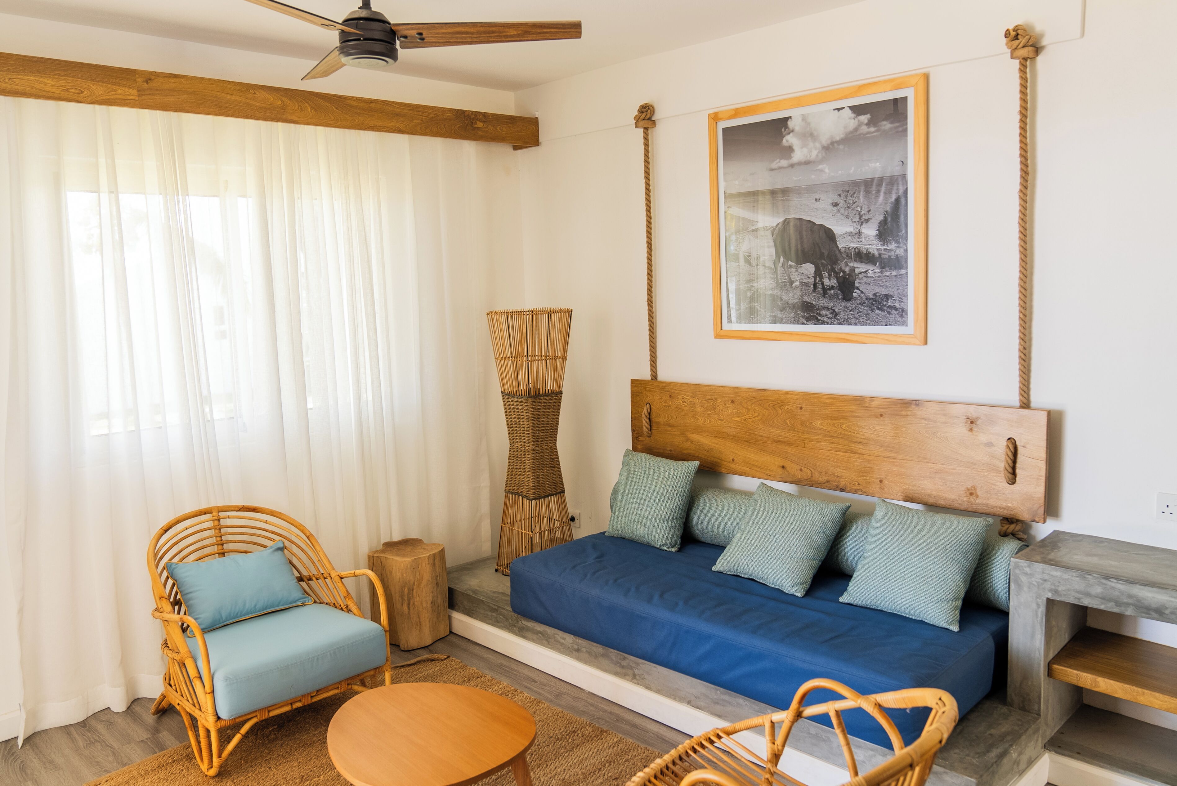 Une pièce avec un canapé bleu, des fauteuils en osier, une tête de lit en bois, un tableau encadré et un ventilateur de plafond.