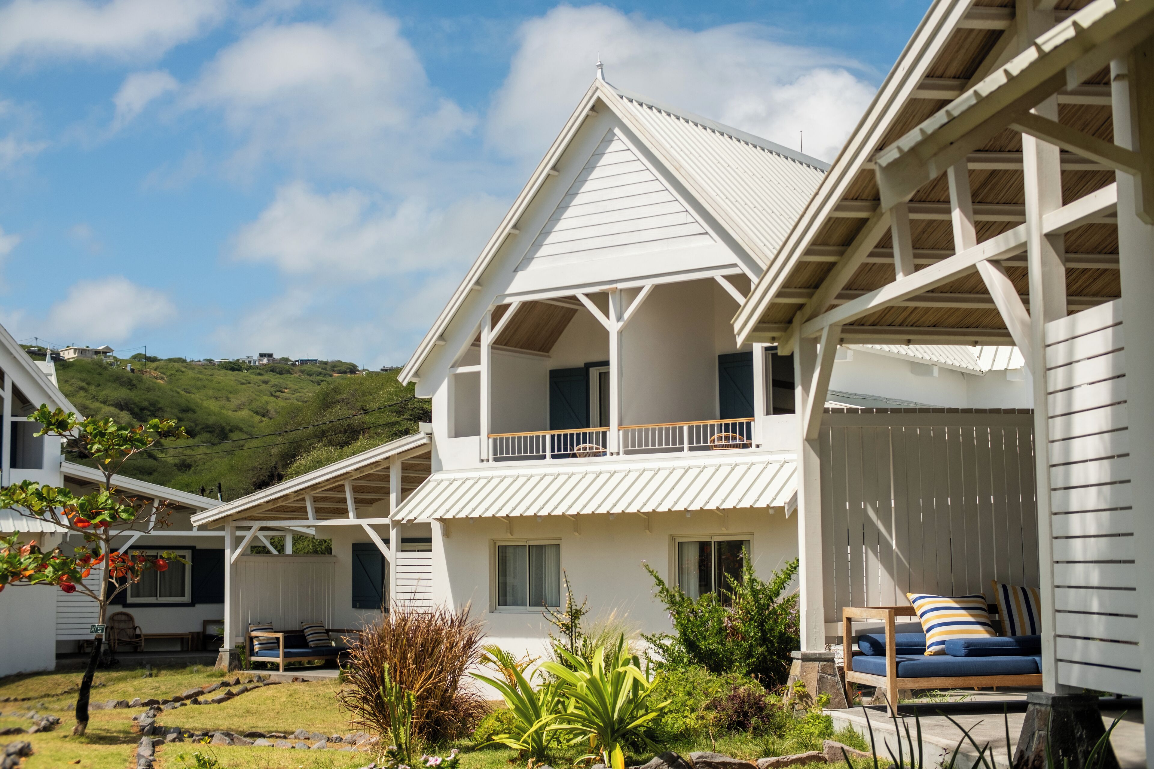 A white two-story house with a balcony, surrounded by greenery and a hilly landscape.
