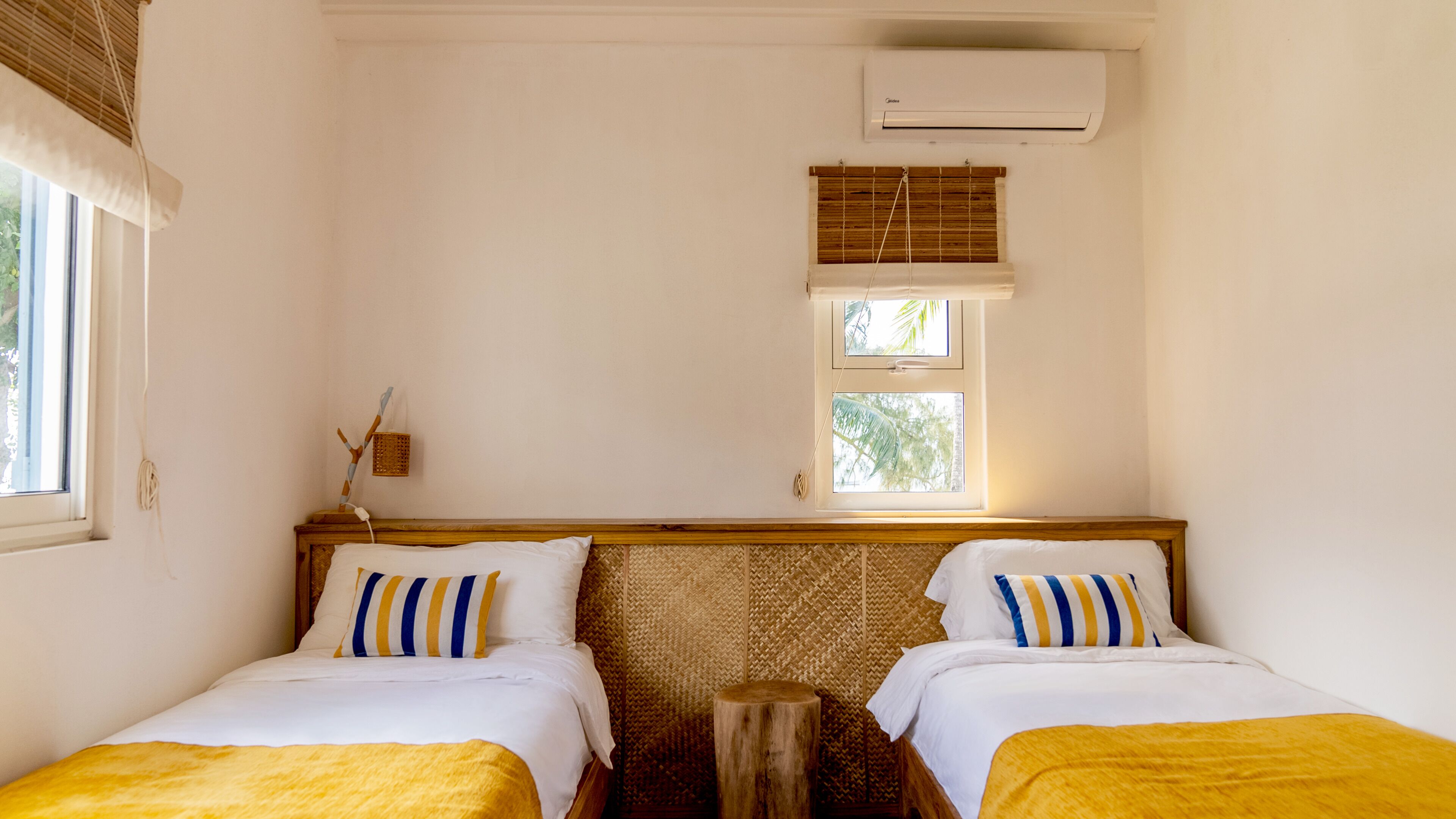 A room with two beds, a window with a view of palm trees, an air conditioning unit, and a woven wall panel.