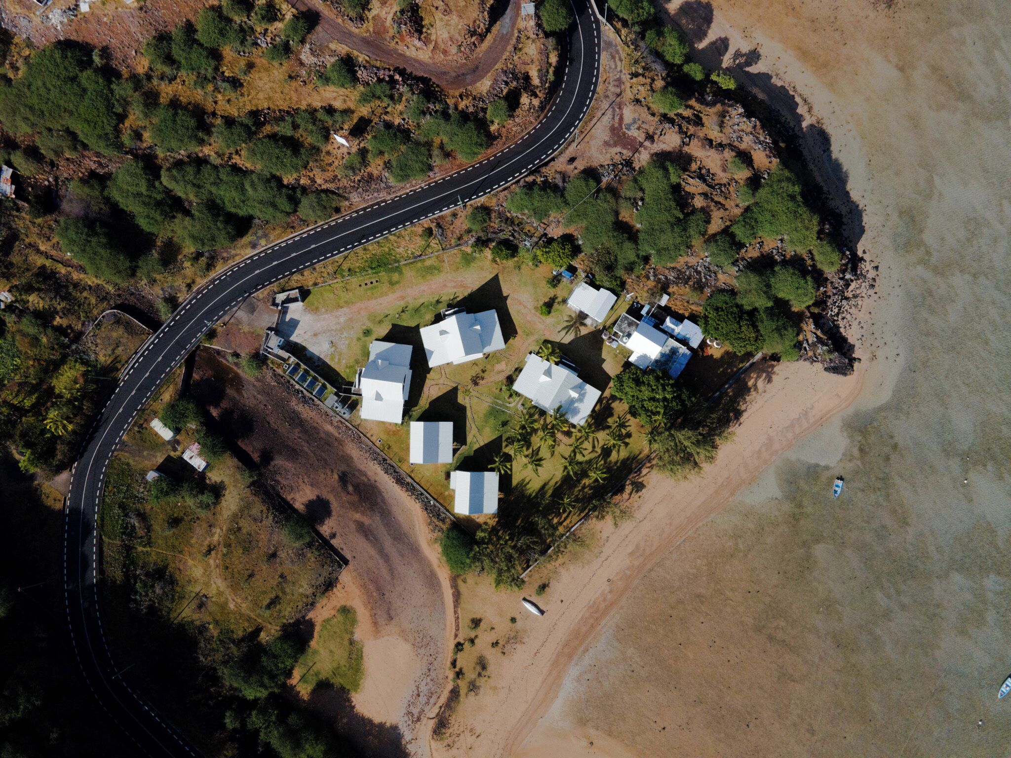 An aerial view of a coastal area with a winding road, several white-roofed buildings, and a beach.