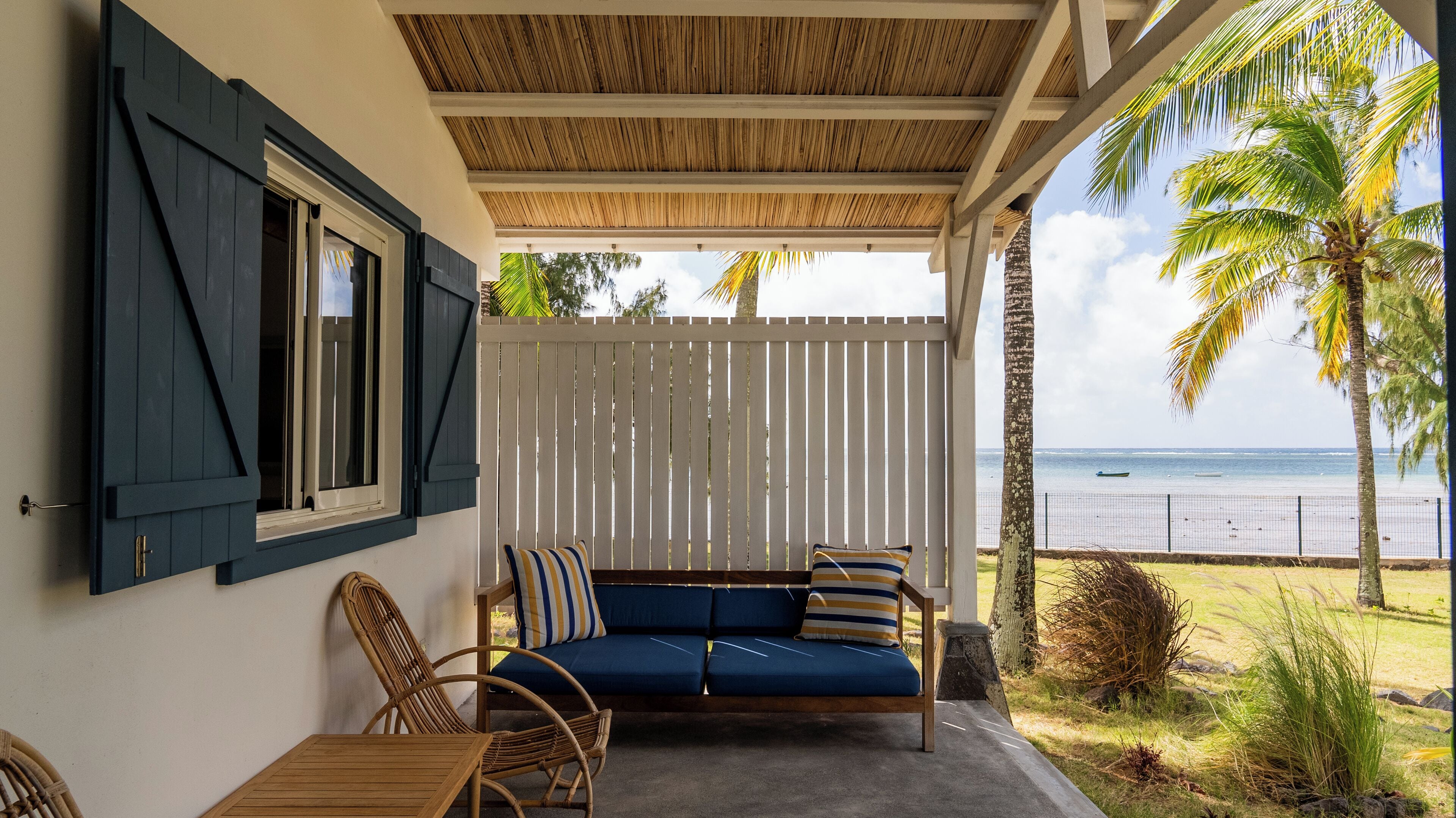 Un espace extérieur aménagé, abrité et doté d’un canapé et de fauteuils, avec vue sur une plage délimitée par une clôture et bordée de palmiers.