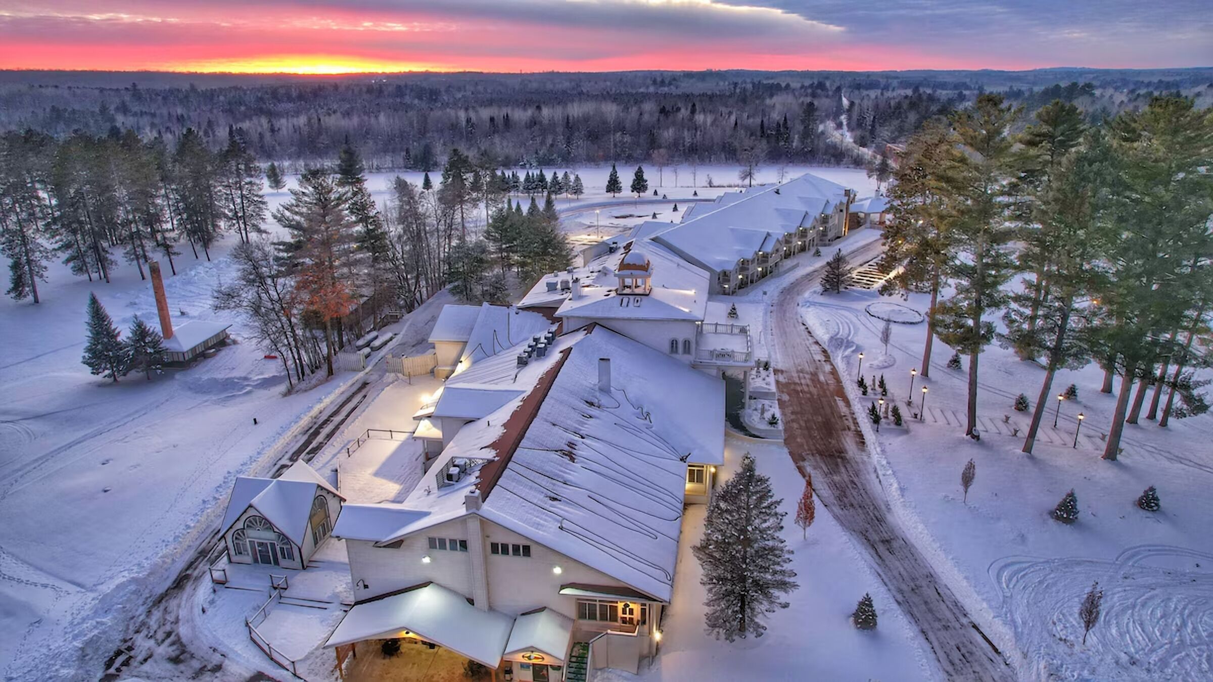 Un grand immeuble à étages, dont le toit est recouvert de neige et qui possède de nombreuses entrées, est entouré d’un paysage enneigé et d’une forêt dense.