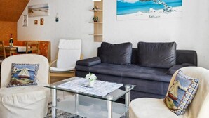 Een moderne woonkamer met een donkergrijze bank, een glazen salontafel en een witte fauteuil. Aan de muur hangt een schilderij van een strandgezicht.