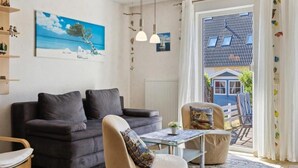 Een woonkamer met een donkere bank, een beige fauteuil, een glazen salontafel en uitzicht op een blauw huis door een groot raam.