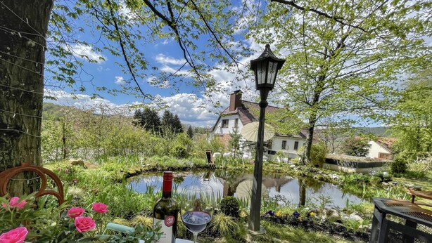 A garden with a pond, a table with a bottle of wine and a glass, and a chair.