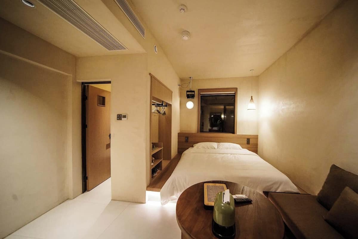 Ein kleines Hotelzimmer mit Bett, Nachttisch, Stuhl und einem Tisch mit einer Flasche.