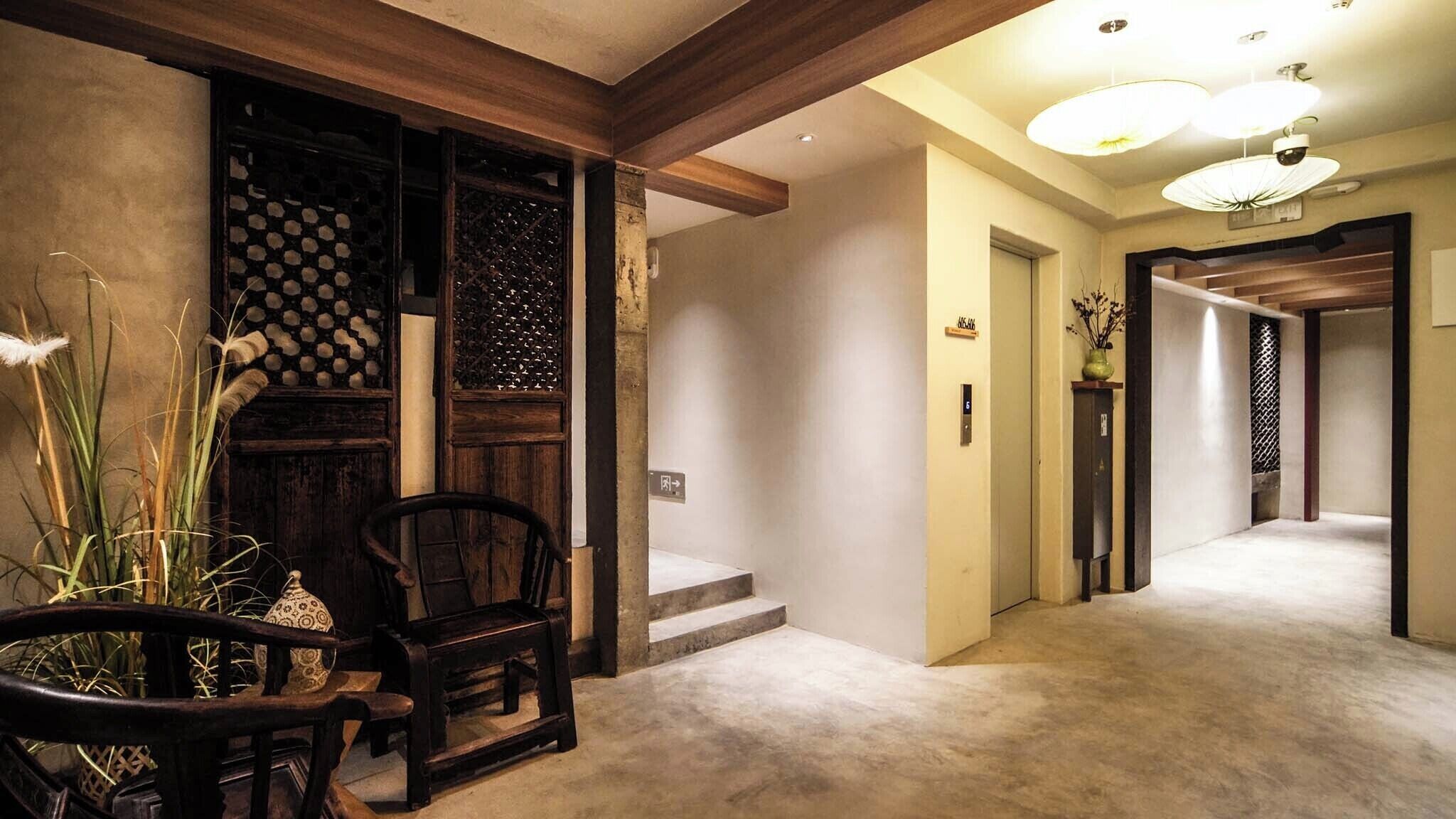 Un couloir avec une chaise en bois, une petite table et un vase décoratif. Des marches mènent à un ascenseur, et une porte est marquée du mot « ASCENSEUR ».