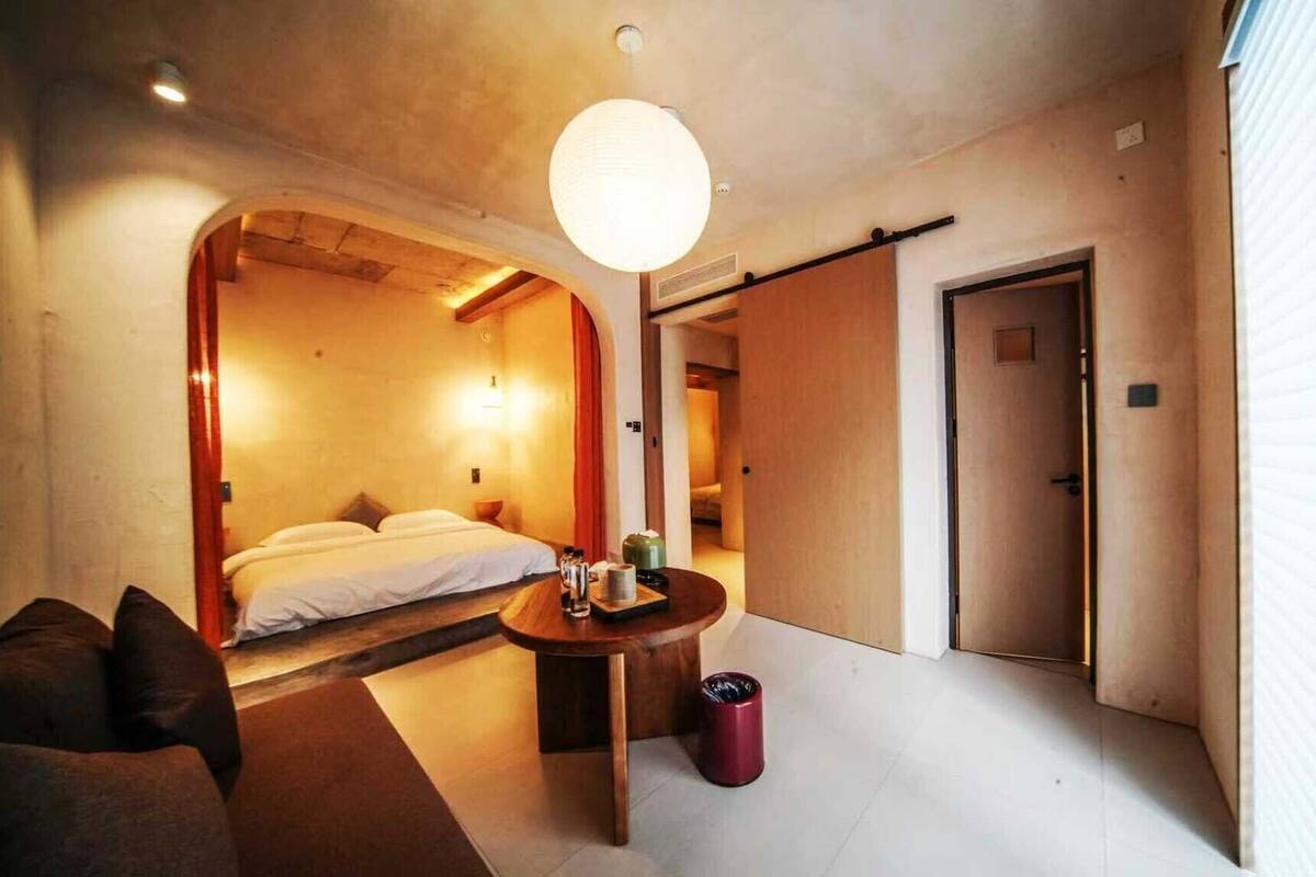 Ein Hotelzimmer mit einem Bett, einem kleinen Tisch mit Lampe, einem Papierkorb und einer Tür zu einem anderen Raum.