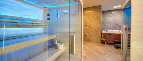 A modern spa room with a glass-enclosed sauna, wooden benches, a massage table, and a small table with a bottle and a bowl.