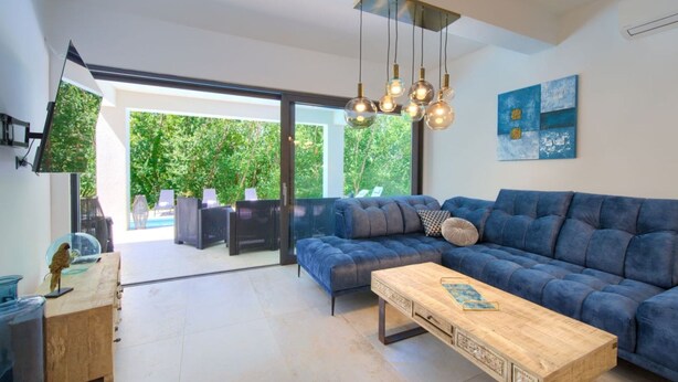 A modern living room with a blue sofa, a wooden coffee table, and a glass-top dining table. There is a large window providing a view of the outdoors. - Wellness Apartment Modruna (Vinodol)