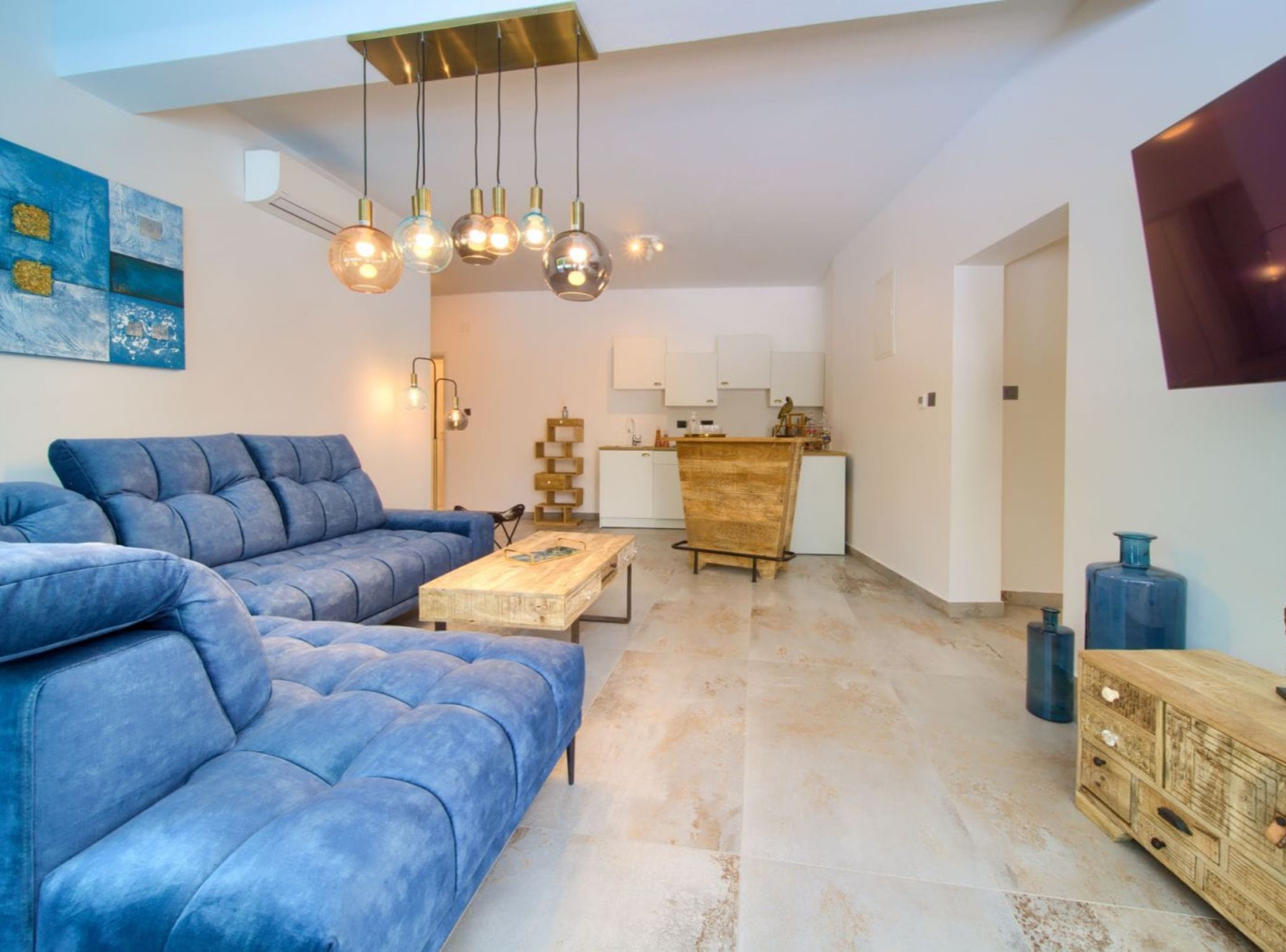 A modern living room with a blue sofa, a wooden coffee table, and a kitchen area with white cabinets and a wooden counter.