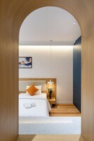Quarto de hotel moderno com chão de madeira, cama com roupa de cama branca e um travesseiro laranja, mesa de cabeceira com um vaso de flores e um quadro na parede.