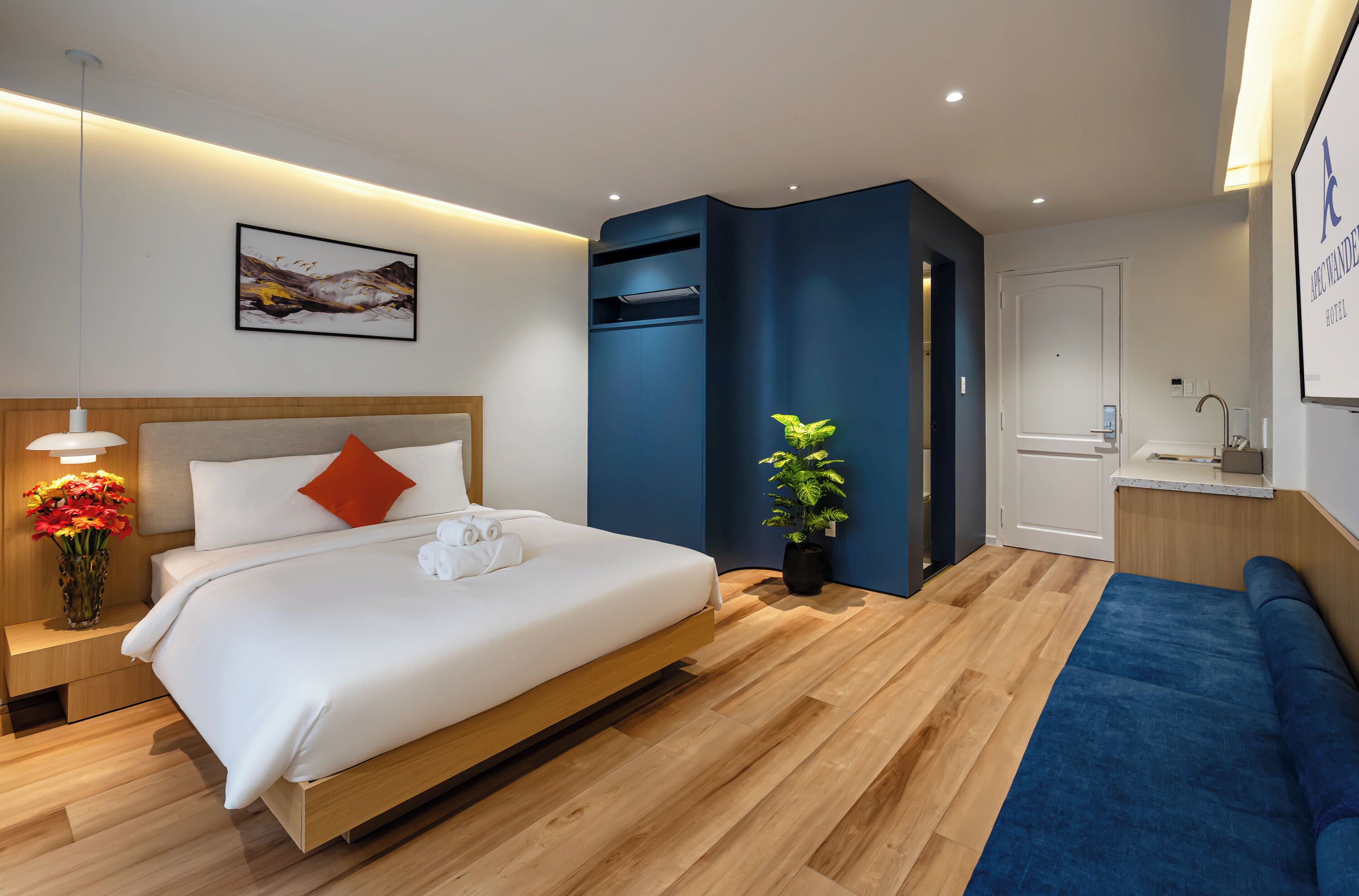 A modern hotel room with a bed, bedside table, lamp, and a potted plant.